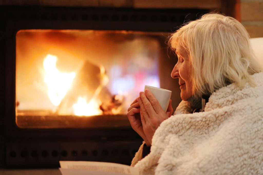 Old lady keeping warm by the fire with a blanket around her and a cup of tea