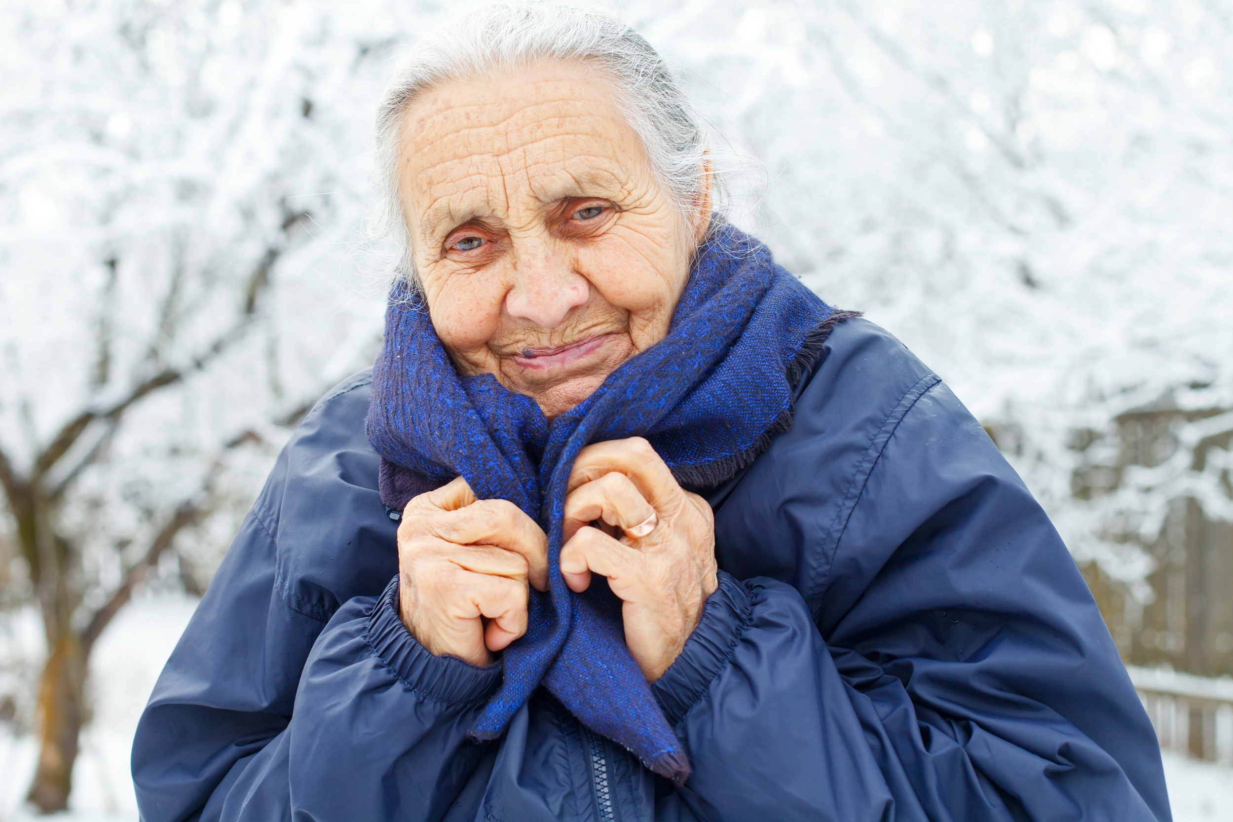 Cold old lady stood outside in snow wearing blue coat and scarf